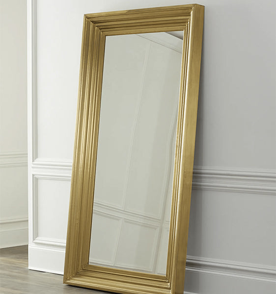 A brass floor mirror with a rectangular frame, placed on a floor against a wall